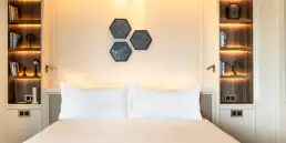Fotografía de interiores profesional enfocada en la zona de cama de una suite en el Hotel Sofitel Barcelona, destacando estanterías con iluminación indirecta y decoración simétrica.