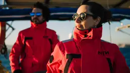 Dos personas vistiendo chaquetas técnicas rojas de la marca NTN Ocean Wear mientras navegan en un velero, con luz dorada de atardecer.